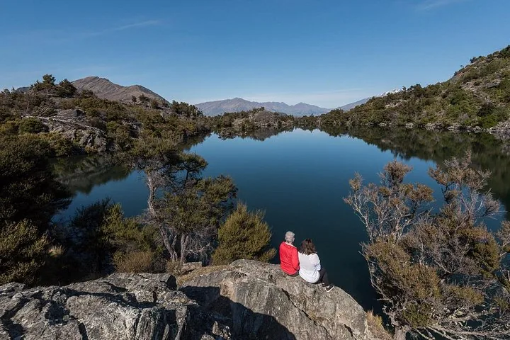 A couple sits on a rock, admiring a calm, reflective mountain lake.