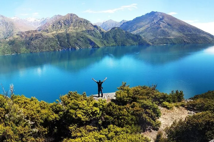 Eine Person steht mit ausgebreiteten Armen auf einer Klippe und blickt auf einen leuchtend blauen See.